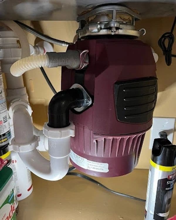 Under-sink garbage disposal unit with plumbing connections and nearby cleaning supplies.