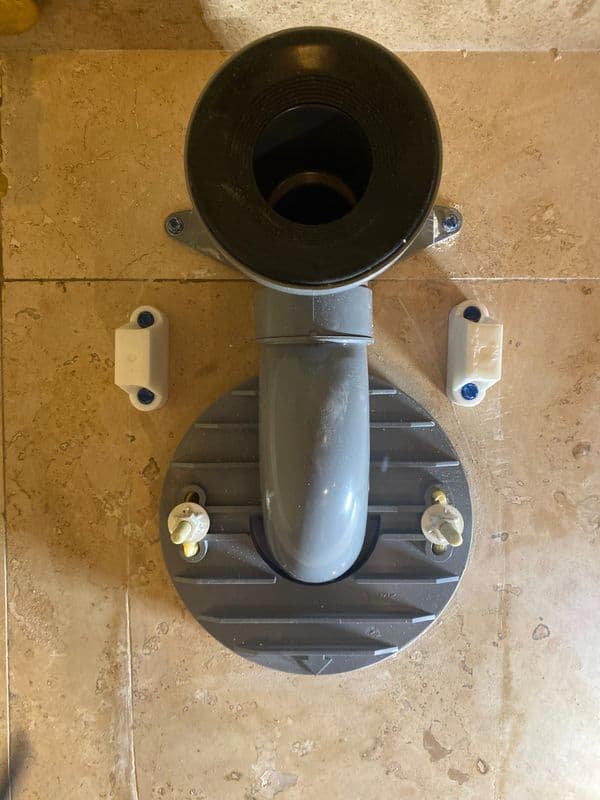 Gray plumbing fixture attached to a tiled floor, featuring water supply connectors and a drain.
