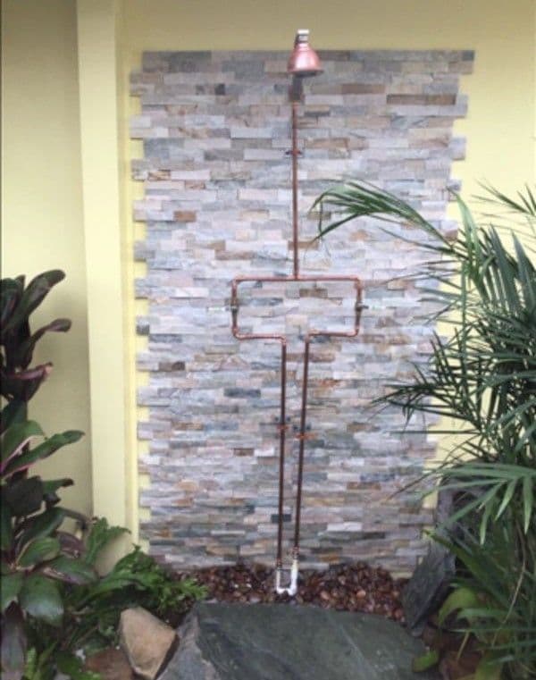 Outdoor shower fixture against a stone wall with tropical plants and decorative stones.