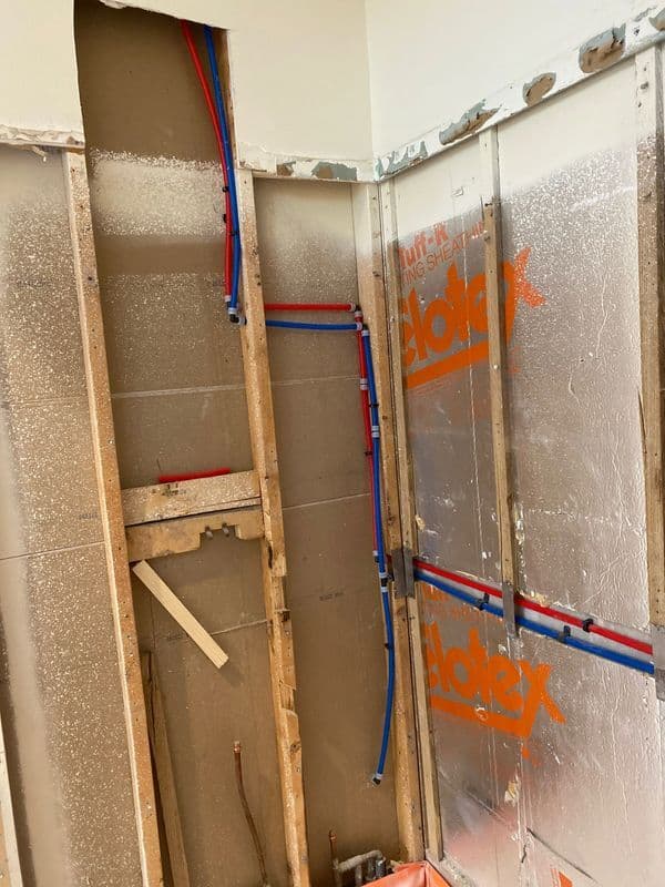 Exposed plumbing with red and blue pipes in a partially renovated bathroom wall.