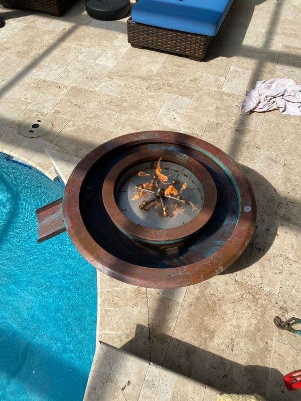 Modern outdoor fire pit surrounded by pool tiles, casting reflections on tranquil water.
