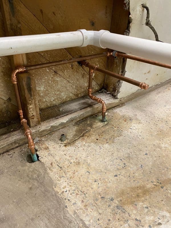 Copper plumbing pipes connected to a wall, with visible water damage on the floor.