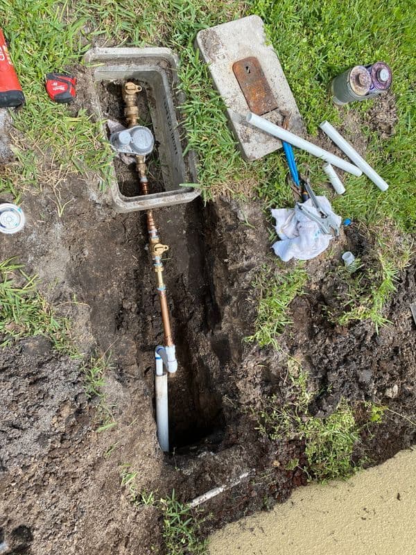 Underground water pipe with valve and tools in a dug trench on grassy area.