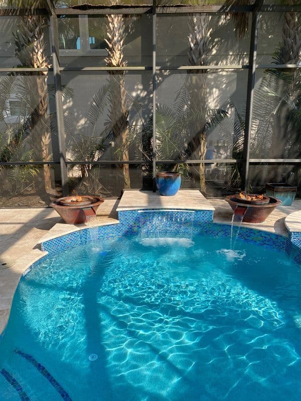 Tropical pool with blue tiles, fountain, and palm trees in a screened enclosure.
