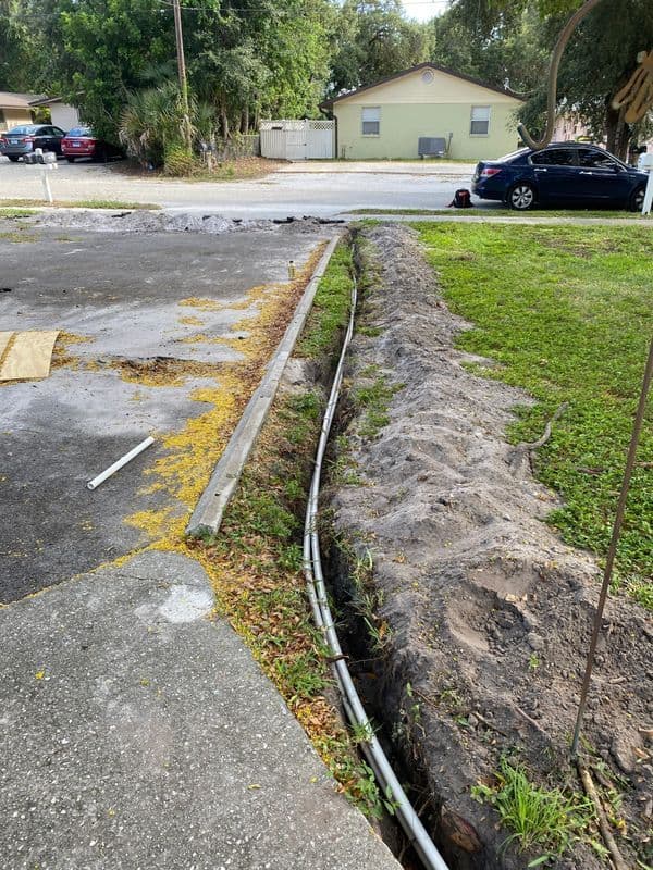 Trench for utilities with exposed pipes in a residential yard, surrounded by grass and pavement.