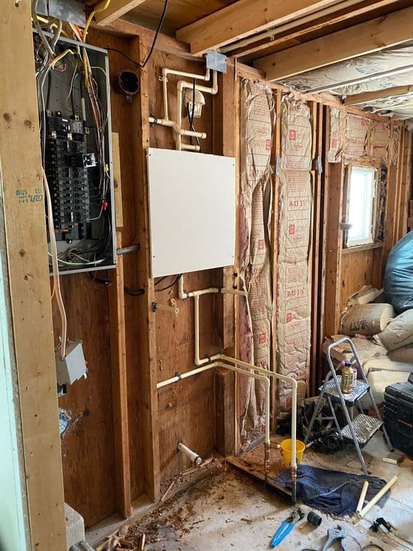 Exposed interior wall with electrical panel, plumbing pipes, and insulation in construction setting.