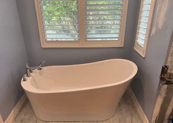 Modern freestanding bathtub in a light blue bathroom with plantation shutters.