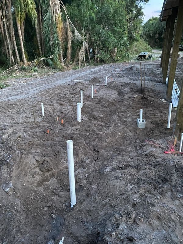 Construction site with PVC pipes installed in muddy ground, surrounded by trees.