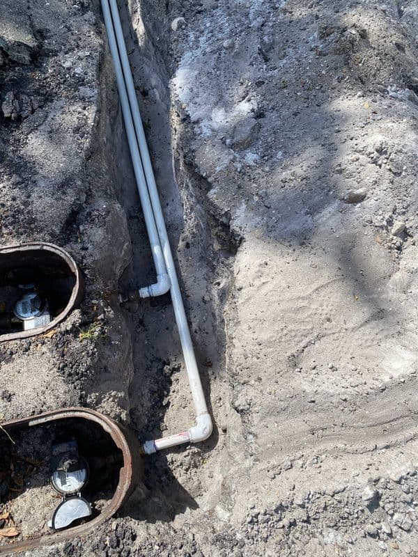 Exposed plumbing pipes in a trench with utility meters and compacted soil.