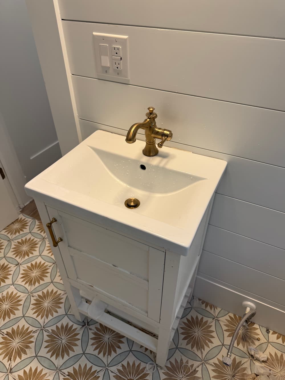 White bathroom sink with brass faucet on patterned tile floor, featuring a wooden cabinet.